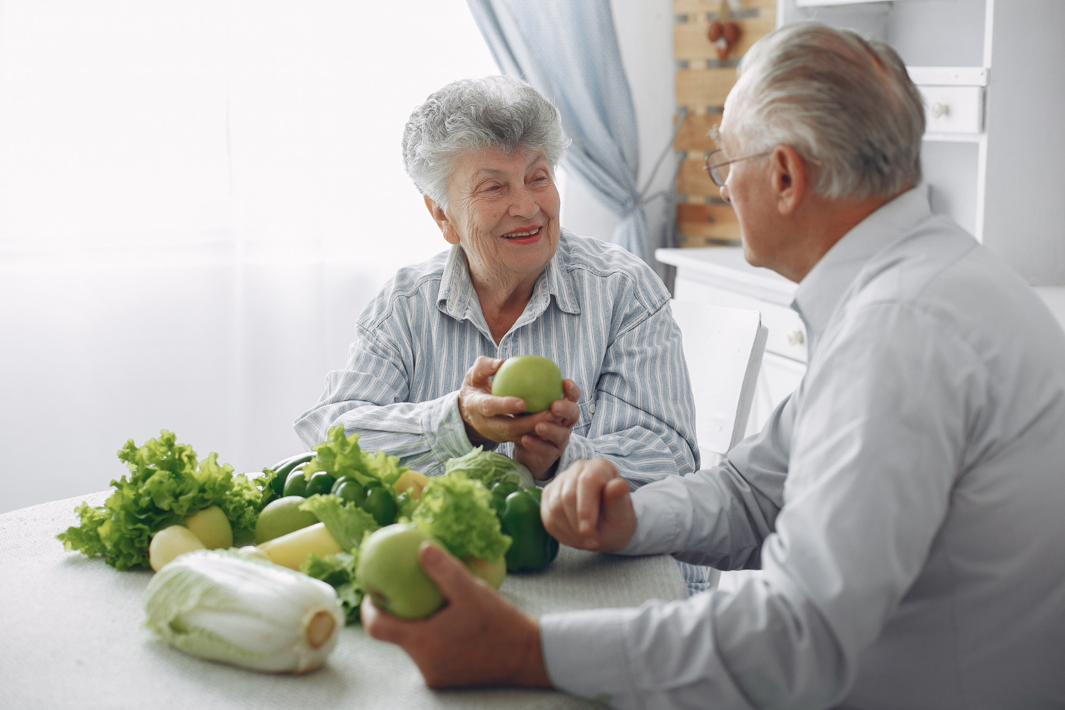 Twee oude mensen met artrose zitten aan een tafel met groenten en fruit die vitamine k bevatten.