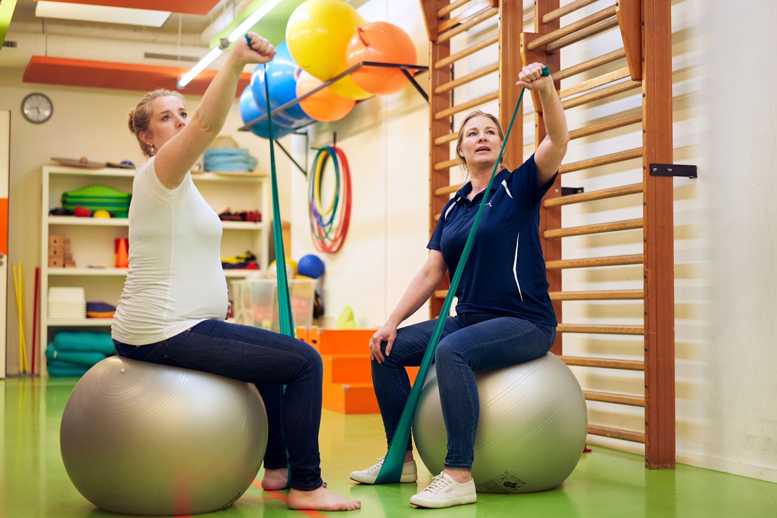 Een fysiotherapeut zit op een fitnessbal in een oefenzaal. Een patiënt zit ook op een fitnessbal. Ze traint met haar eigen lichaamsgewicht. Ze stretchen allebei hun armen.